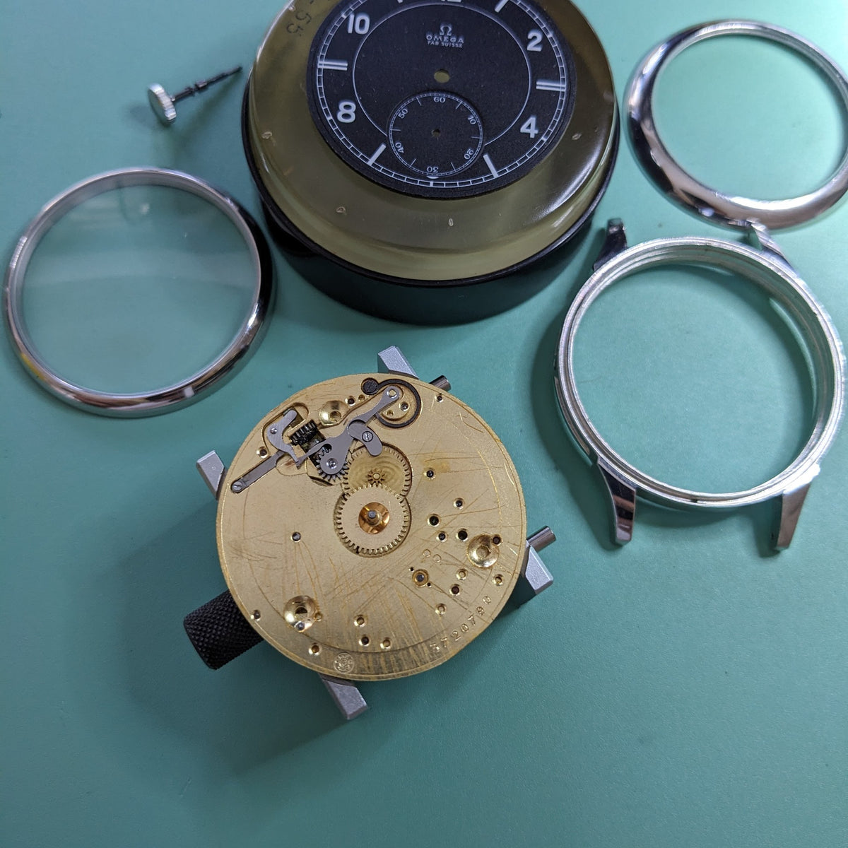 Omega-Uhrkomponenten auf einer türkisfarbenen Oberfläche, einschließlich des dunklen Zifferblatts, freiliegendem mechanischen Uhrwerk, Gehäuseringen, Glas und