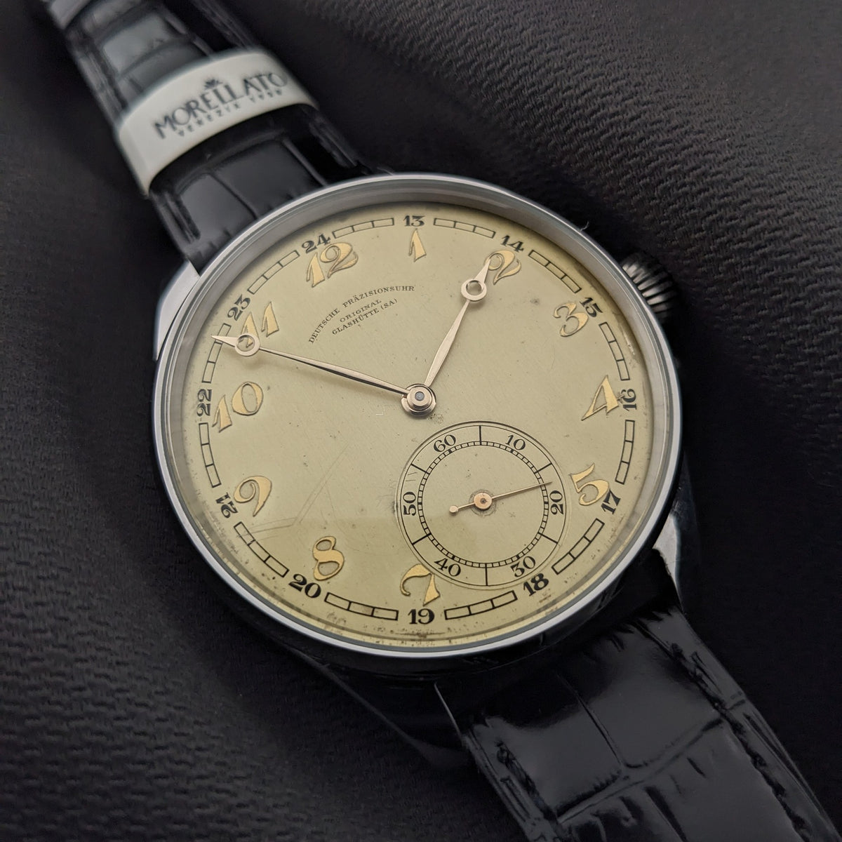 Deutsche Präzisions-Uhrenfabrik Glashütte Uhr auf dunklem Stoff, mit cremefarbenem Zifferblatt, schwarzem Krokodilmusterarmband und Armbandetikett.