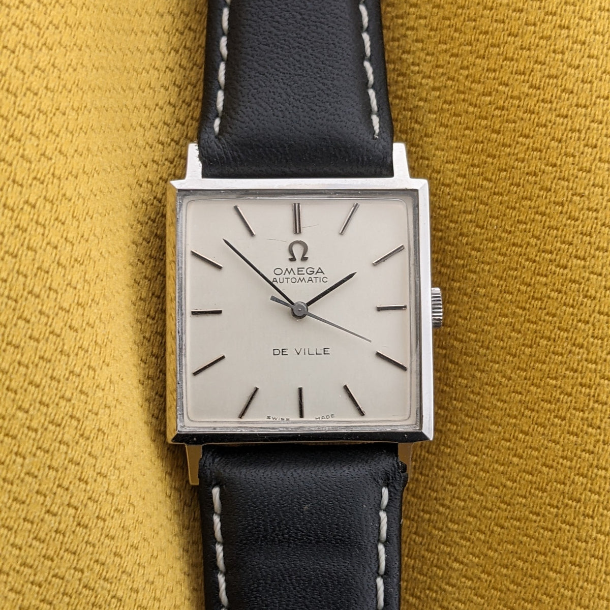 Frontansicht der Omega De Ville Automatik 161.014 auf senffarbenem Stoff, zeigt helles quadratisches Zifferblatt, Stabindizes und schwarzes Armband.