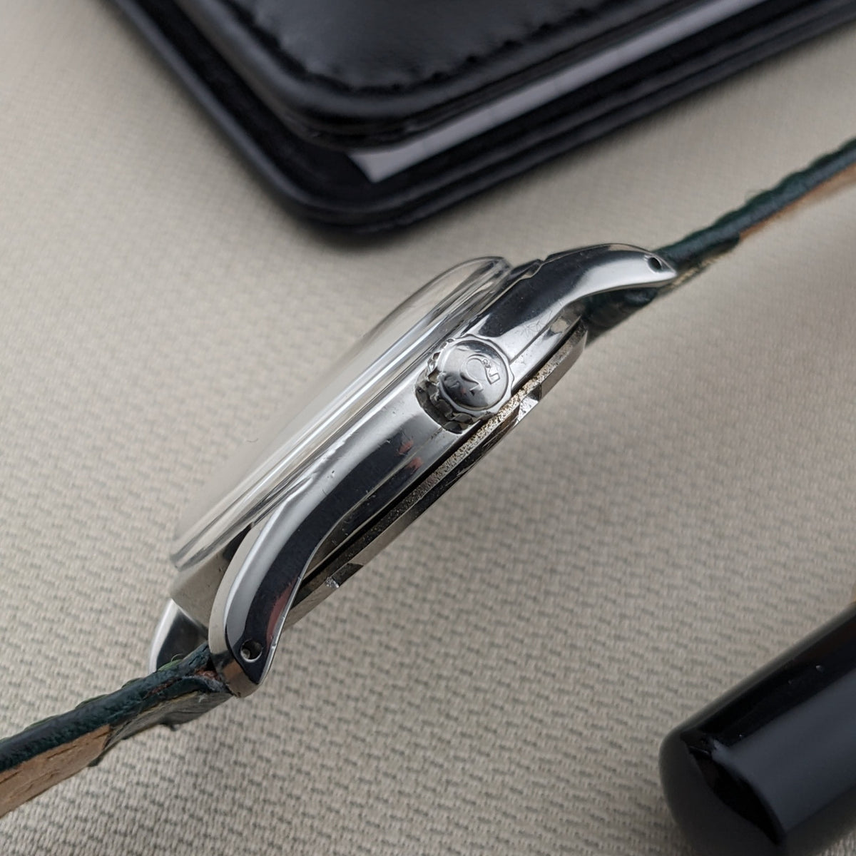 Profilo laterale dell'OMEGA Classic referenza 2639-11 con cassa lucida, corona, anse curve, cinturino verde e cassa nera sullo sfondo.