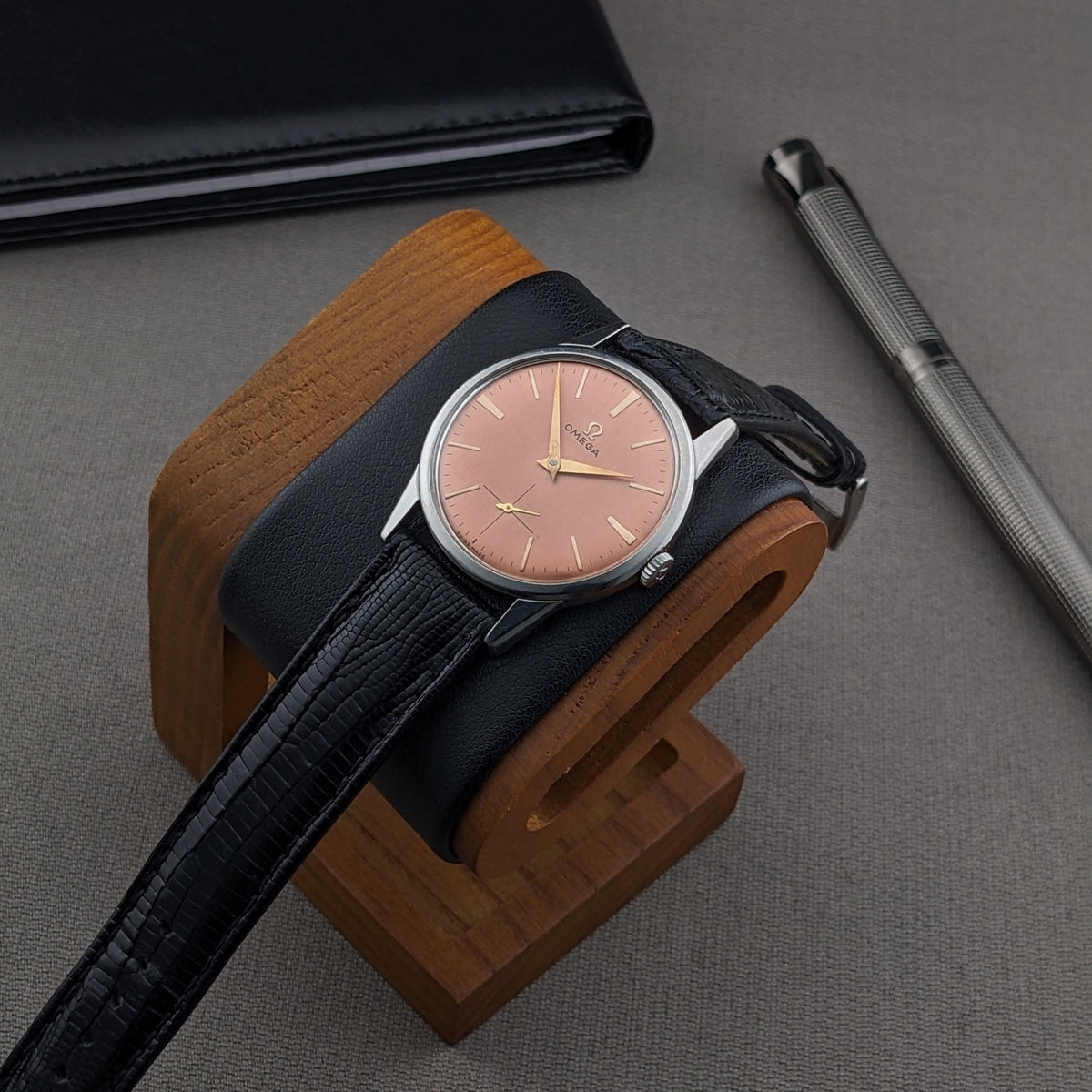 Omega Classic referenza 14391-1 su supporto in legno con cuscino nero, cinturino nero, portafoglio e penna sullo sfondo.