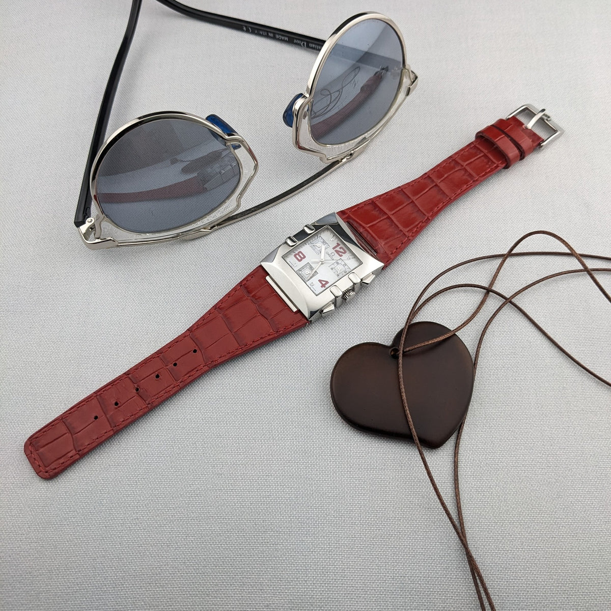 Omega Constellation Quadra 1841.73.31 arranged with round sunglasses, a heart-shaped pendant, and its red crocodile-pattern strap.