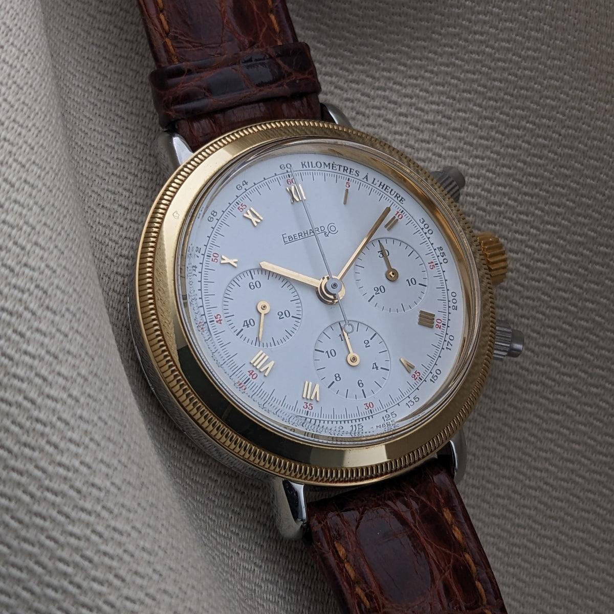 Eberhard & Co.. Godron Chrono reference 32031 lies on textured fabric, displaying its fluted gold-tone bezel, white dial and crocodile-pattern strap.