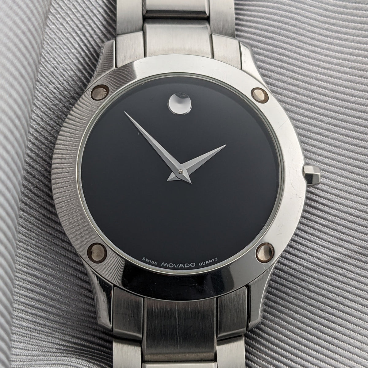 Vue de face d'une montre Movado avec cadran minimaliste noir, aiguilles argentées, repère circulaire et bracelet en acier inoxydable.