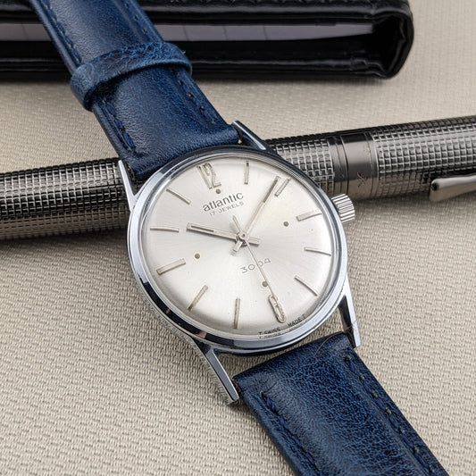 Atlantic 3004 angled across a textured surface with a silver dial, blue leather strap, polished case, and a metal pen in the background.