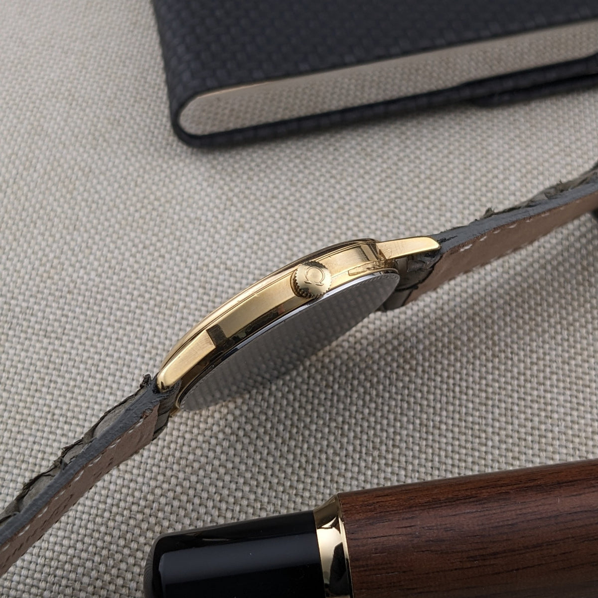 Side view of the gold-tone Omega De Ville reference 111 0156 case and crown, with the strap crossing a woven surface.