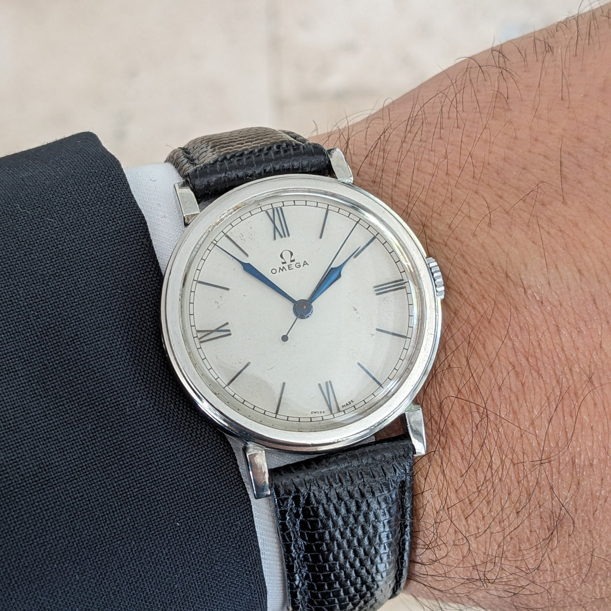 Omega watch worn on a wrist, showing a light dial with Roman numerals, blue hands and a black textured leather strap.