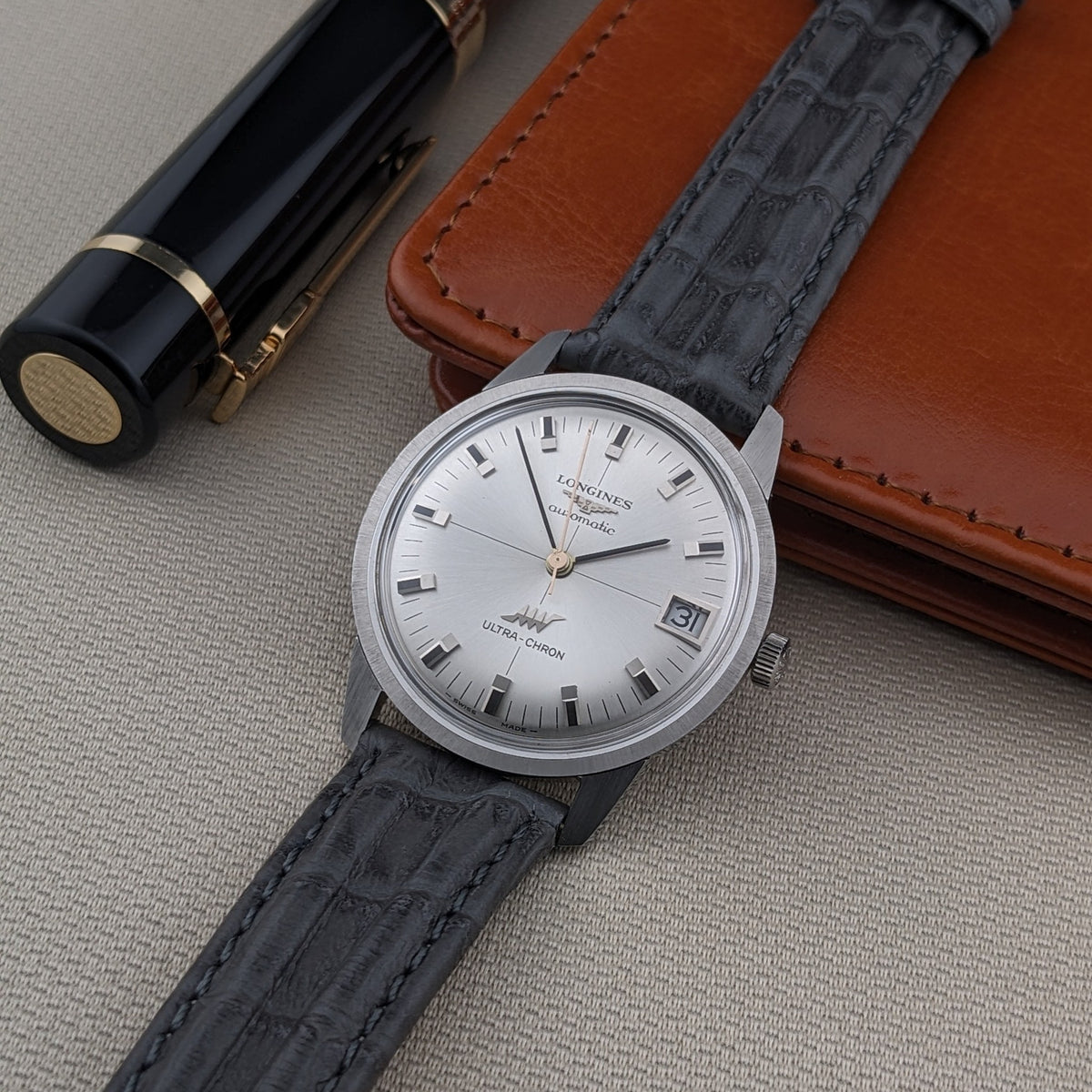Longines Ultra-Chron 7827 on a black leather strap, showing its silver dial, date window, and baton markers beside a pen and leather case.