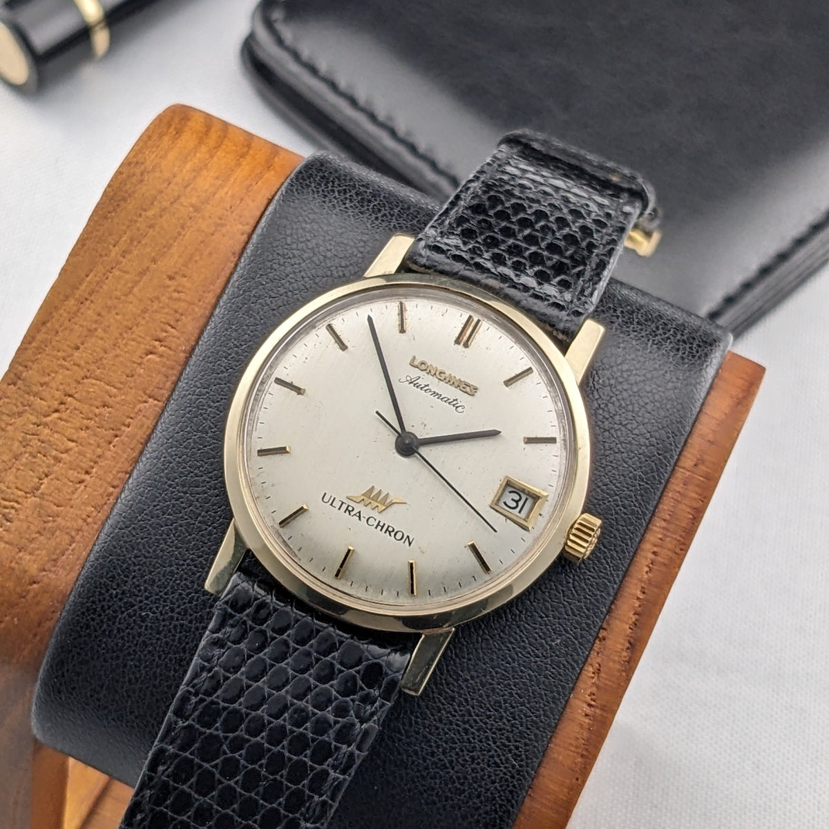 Longines Ultra-Chron 69-556AC centered on a black cushion with cream dial, gold-tone case, date window, and black textured strap.