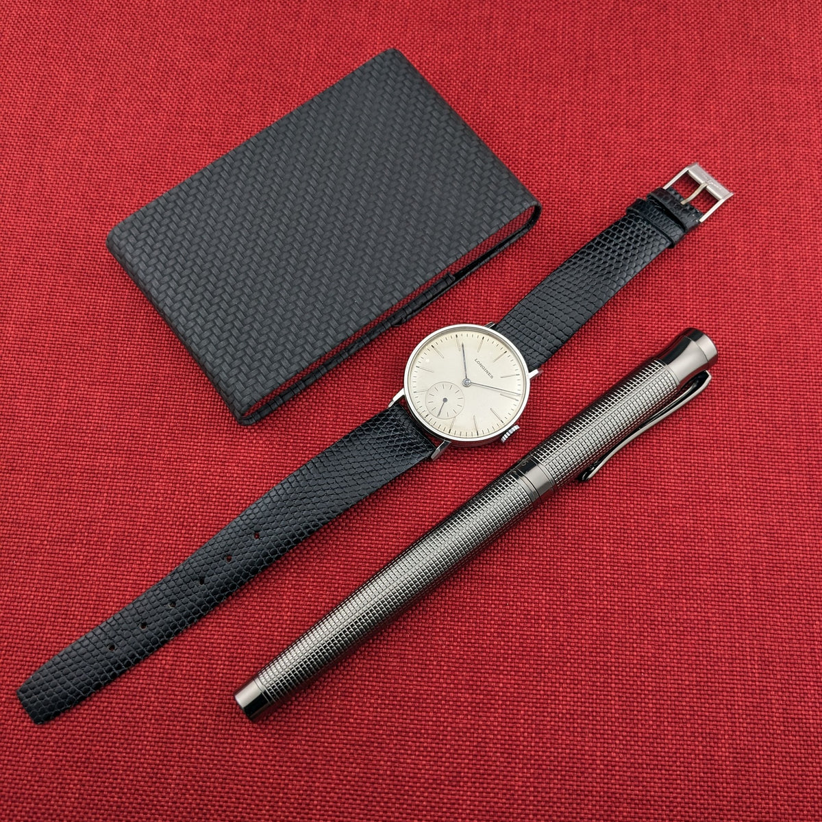 Longines Calatrava reference 20757 arranged on red fabric with a black textured notebook and a silver patterned pen.