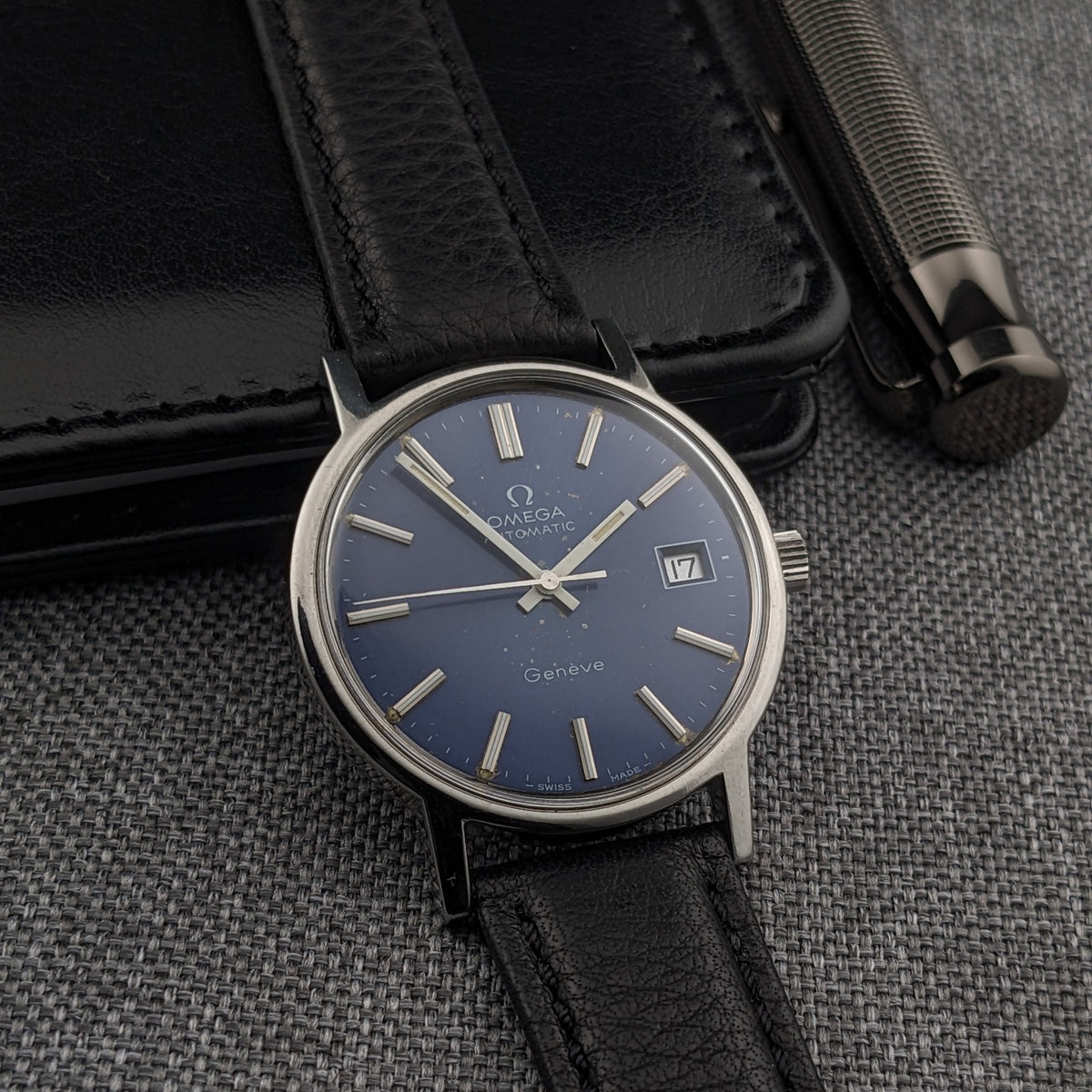 Omega Geneve Automatic reference 166.0163 with blue dial, applied hour markers, date window and black leather strap beside a black case.