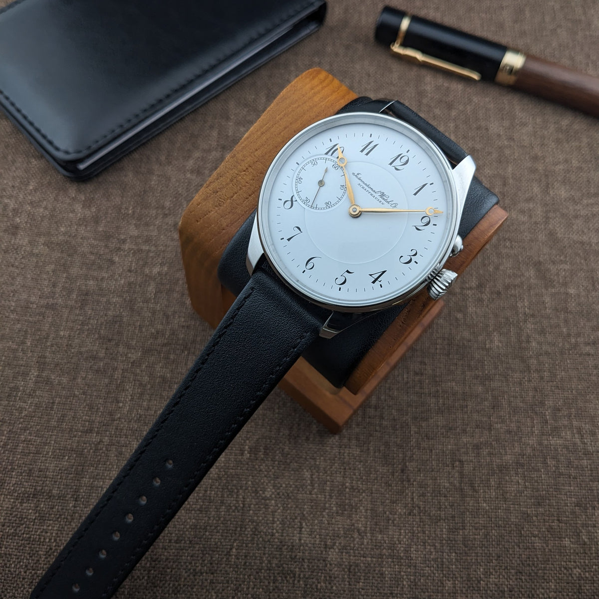 IWC Schaffhausen घड़ी का लकड़ी के स्टैंड पर कोणीय दृश्य, सफेद डायल, सुनहरे हाथ और काले पट्टा के साथ।