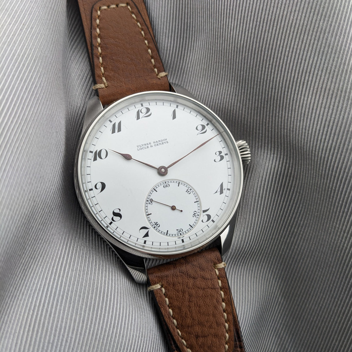 Ulysse Nardin कलाई घड़ी का कोणीय दृश्य, जिसमें सफेद अंक डायल, छोटे सेकंड रजिस्टर और भूरा सिला हुआ चमड़े का पट्टा दिखता है।
