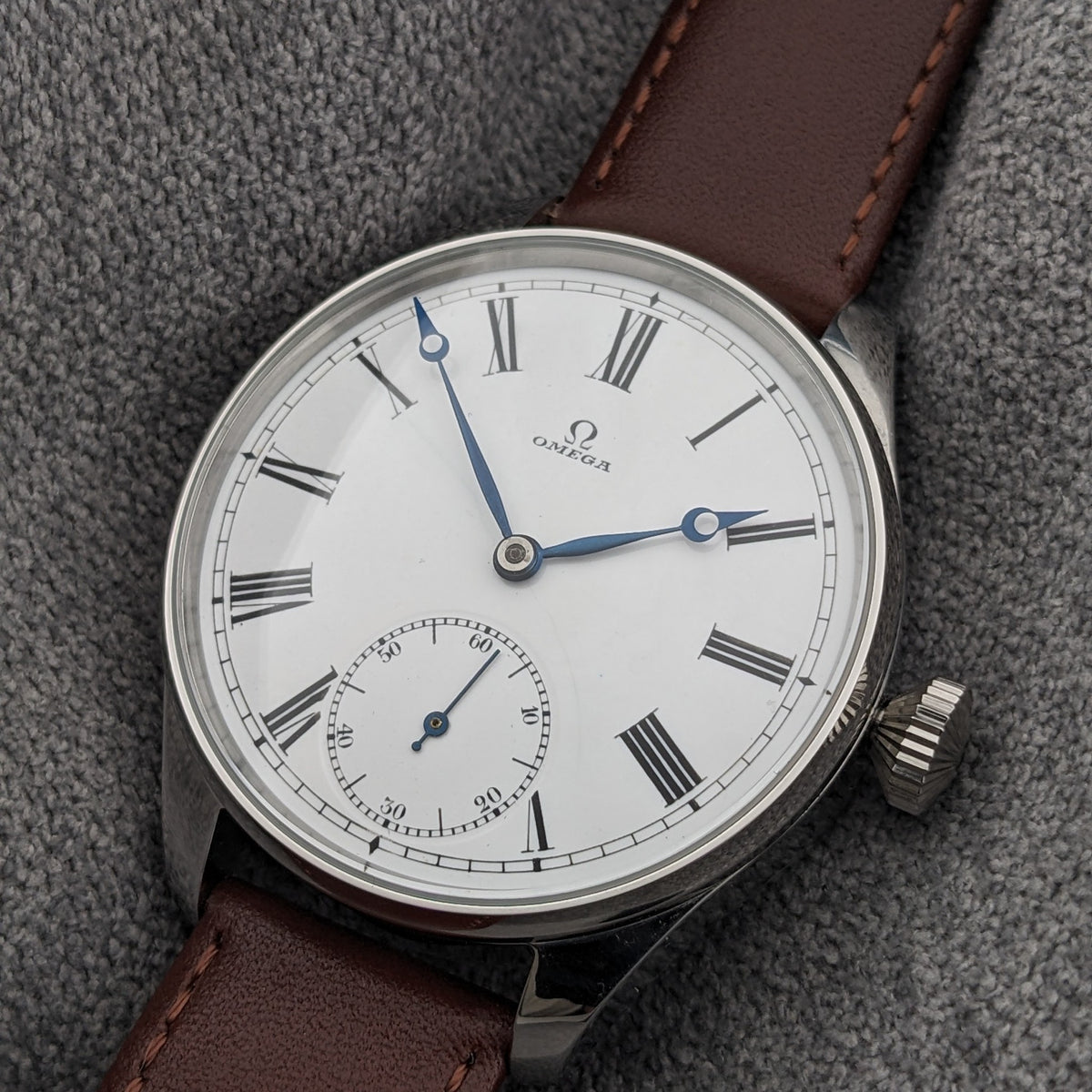 Orologio Omega su tessuto grigio, con quadrante bianco, numeri romani neri, lancette blu, piccolo quadrante dei secondi e cinturino in pelle marrone.