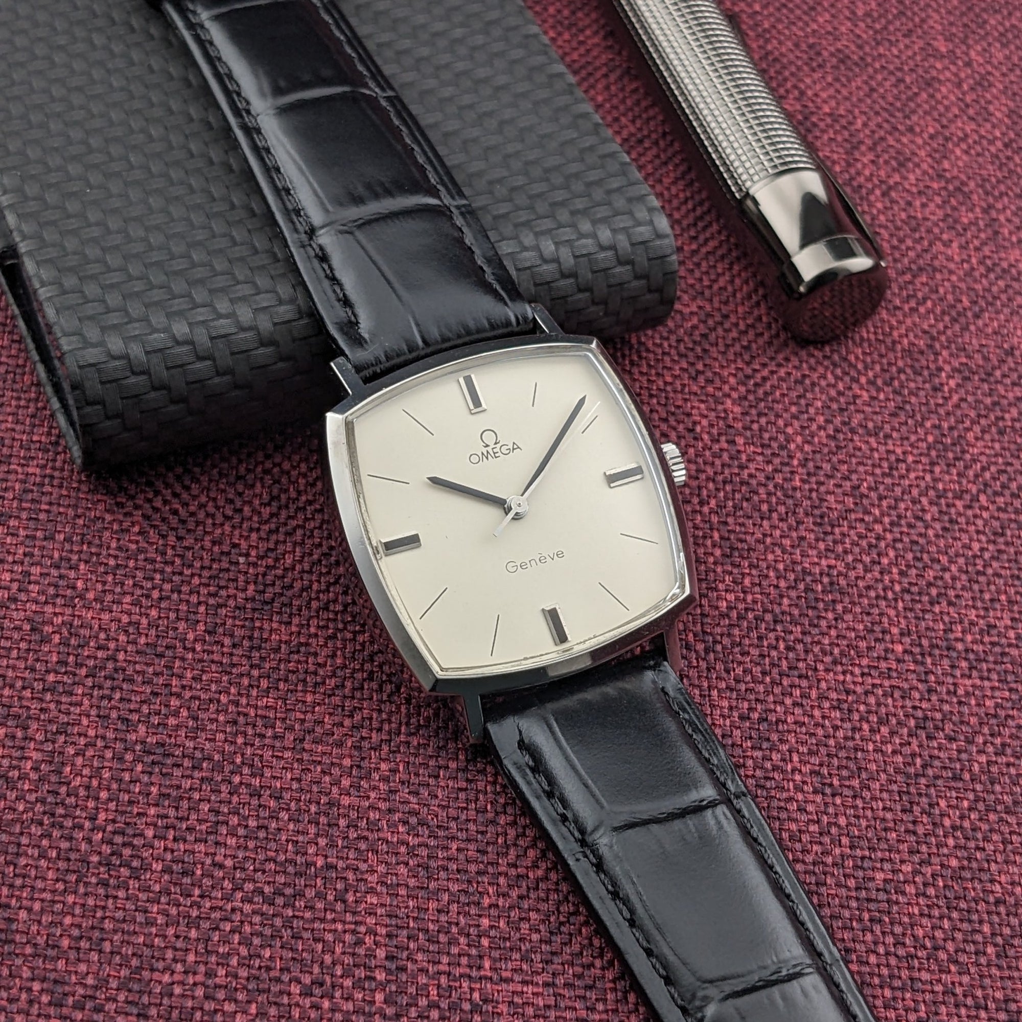 Omega Genève reference 131.022 shown vertically beside a dark textured case and silver pen, highlighting the square cream dial.