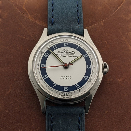 Atlantic Varldsmastarur displayed vertically on a brown leather cover, showing the white dial, navy hour ring, and blue stitched strap.