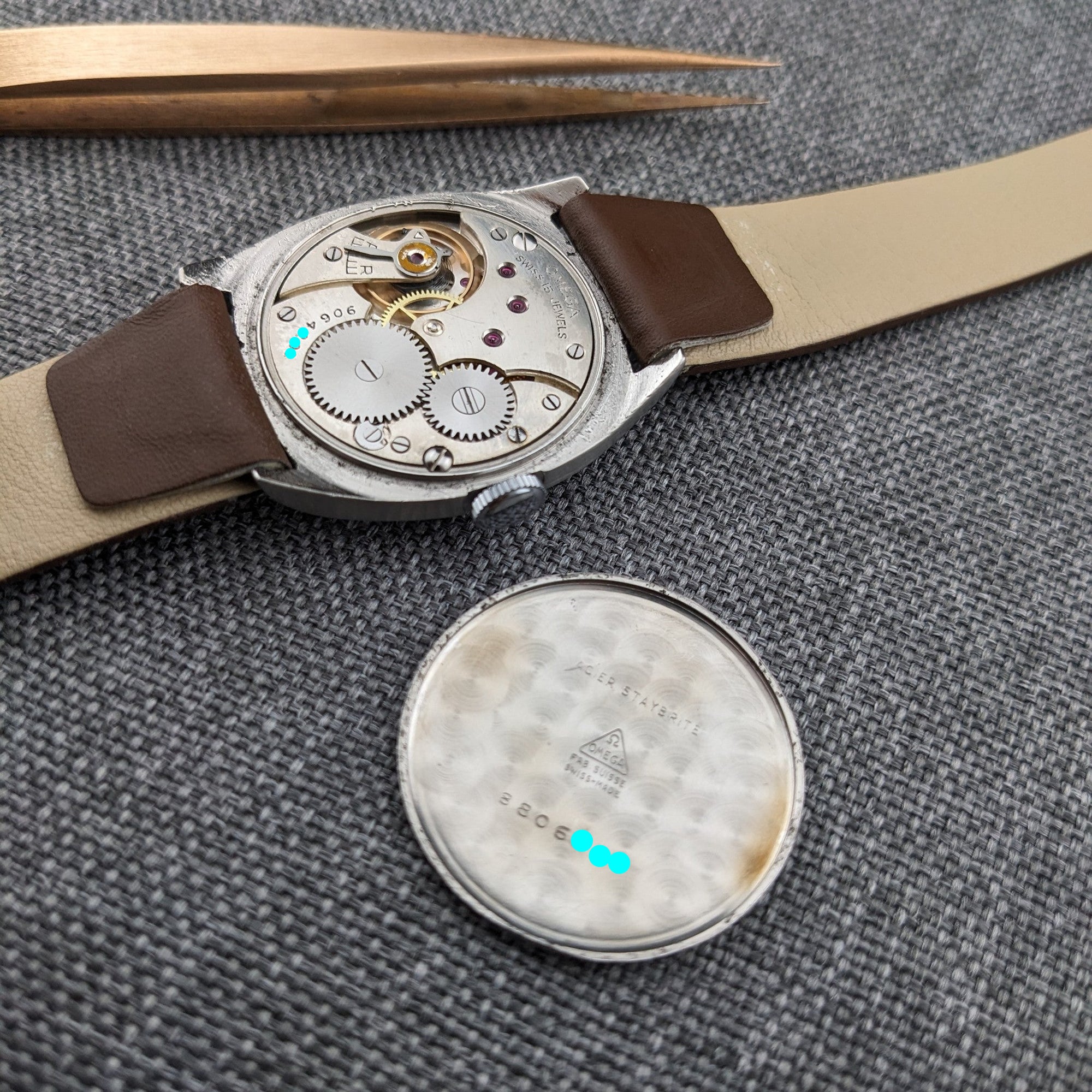 Omega mechanical movement and separate caseback displayed beside the brown leather strap on gray woven fabric, with tweezers above.