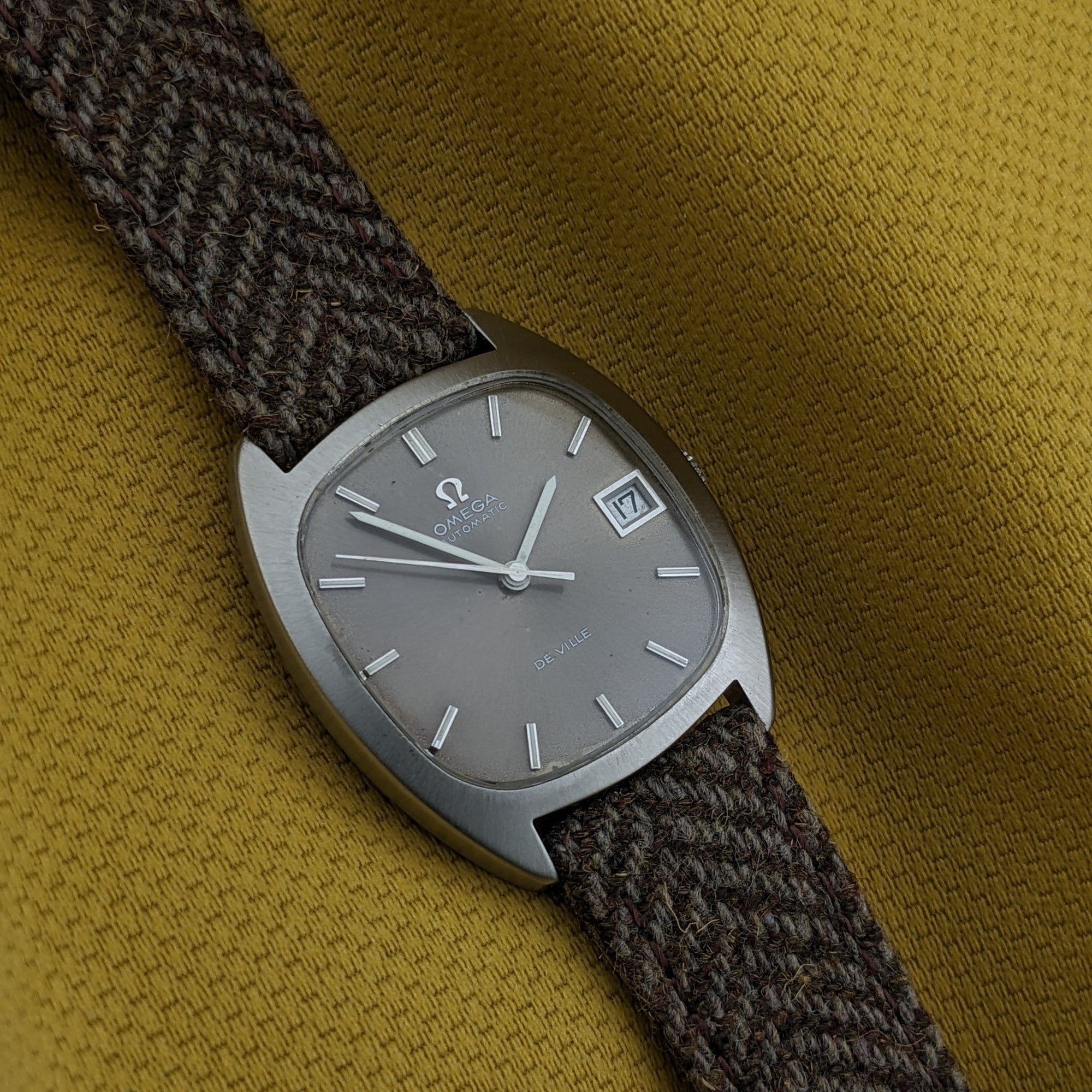 Close-up of the Omega Dr Ville Automatic ref. 162.045 dial on yellow fabric, featuring baton markers, date window, and brown woven strap.