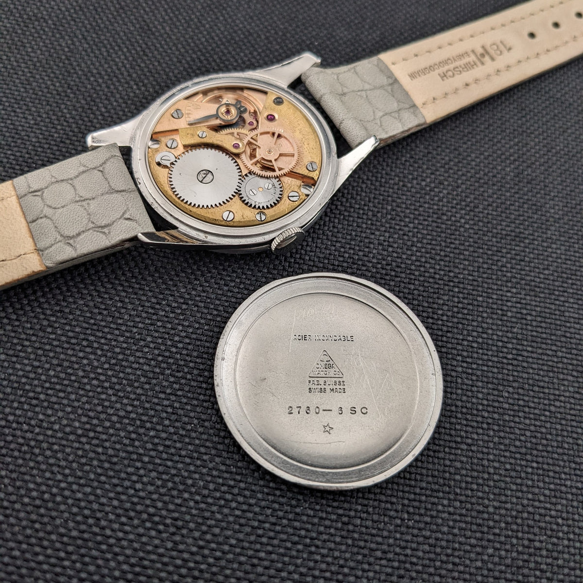 Omega Classic 2760-6 SC mit freigelegtem Werk neben abgenommenem Gehäuseboden, dessen Innengravur auf schwarzer Textur sichtbar ist.