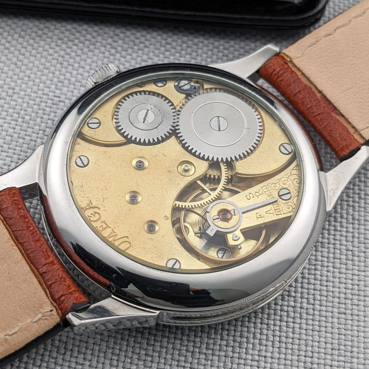 Omega-Armbanduhr durch den transparenten Sichtboden betrachtet, zeigt Zahnräder und das mechanische Uhrwerk zwischen braunen Lederarmbandteilen.