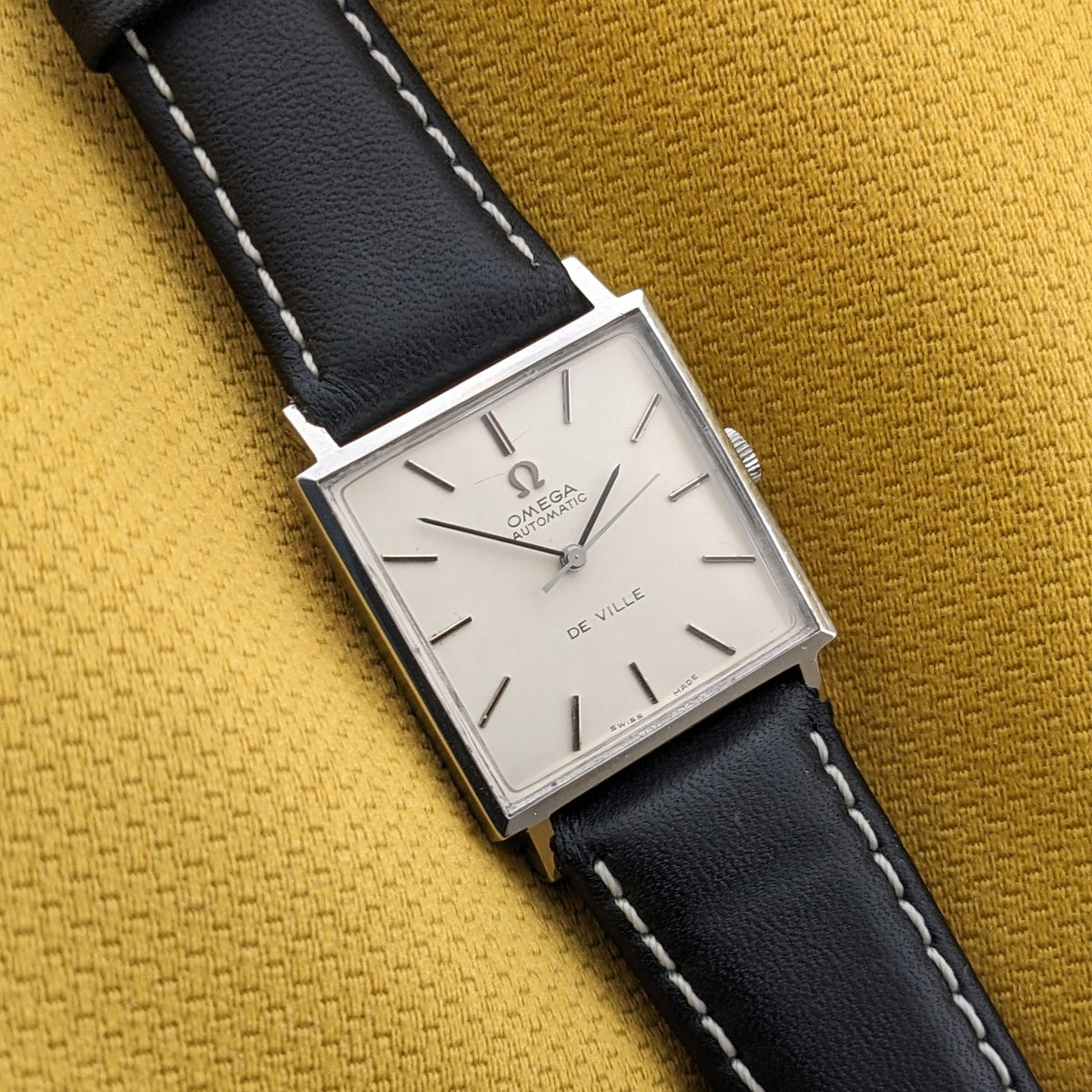 Schrägansicht der Omega De Ville Automatik 161.014, mit hellem rechteckigem Zifferblatt und schwarzem Lederarmband auf strukturiertem senffarbenem Stoff.