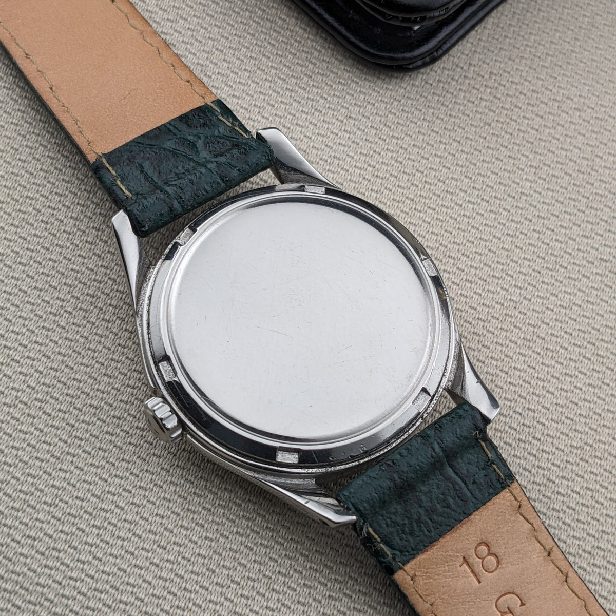 Vista posteriore dell'OMEGA Classic 2639-11 con fondello circolare liscio, cinturino verde, fodera color cuoio e custodia in pelle nera dietro.