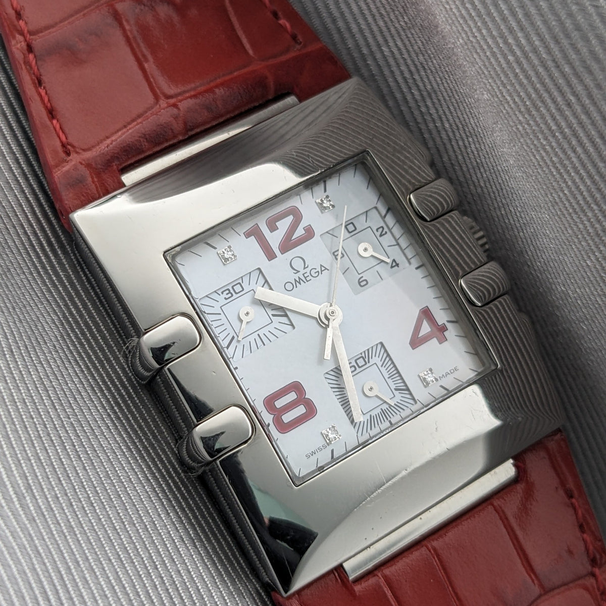 Angled close-up of the Omega Constellation Quadra 1841.73.31, showing its white dial, red numerals, polished rectangular case, and red strap.