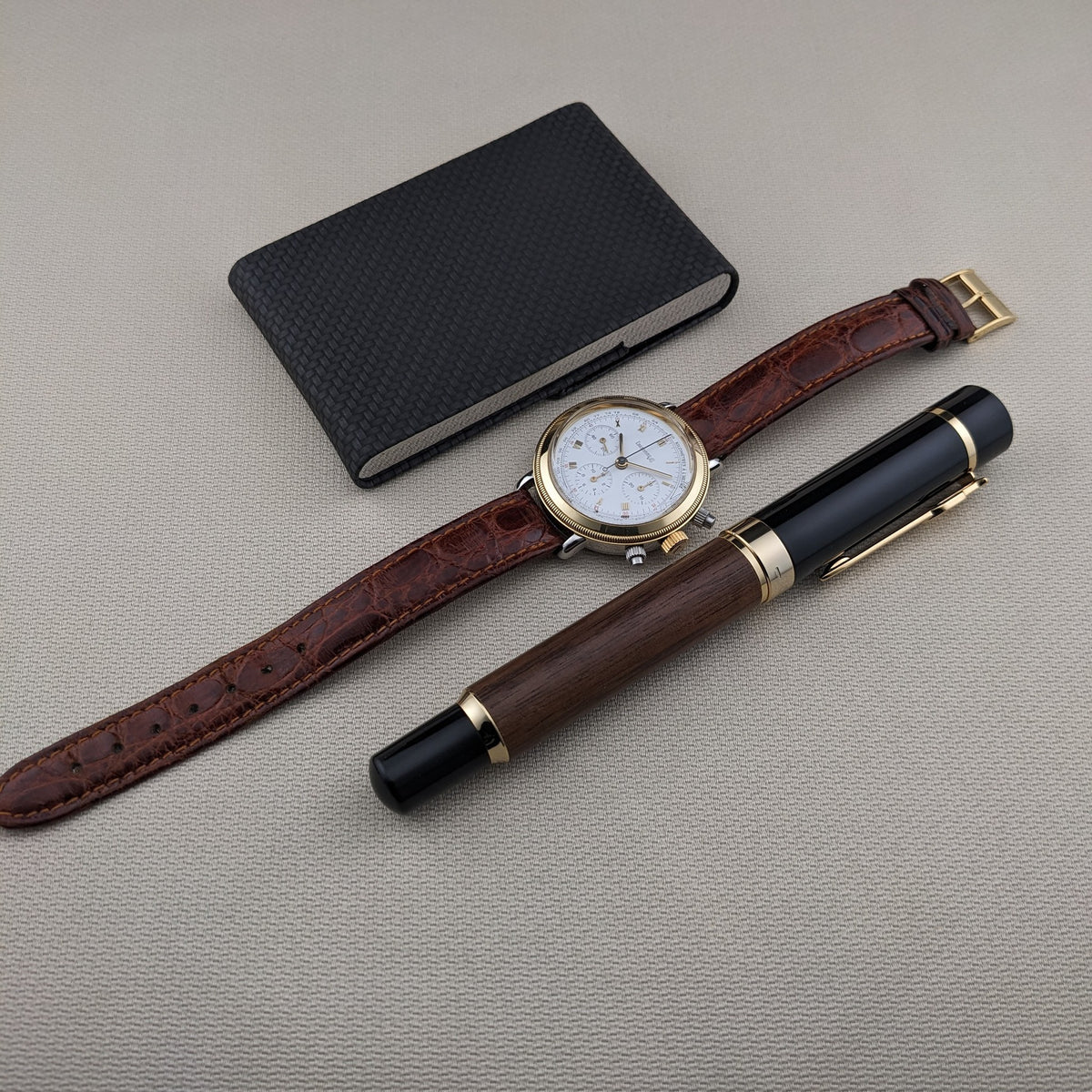 Eberhard & Co.. Godron Chrono reference 32031 is arranged diagonally beside a black notebook and two fountain pens on light fabric.