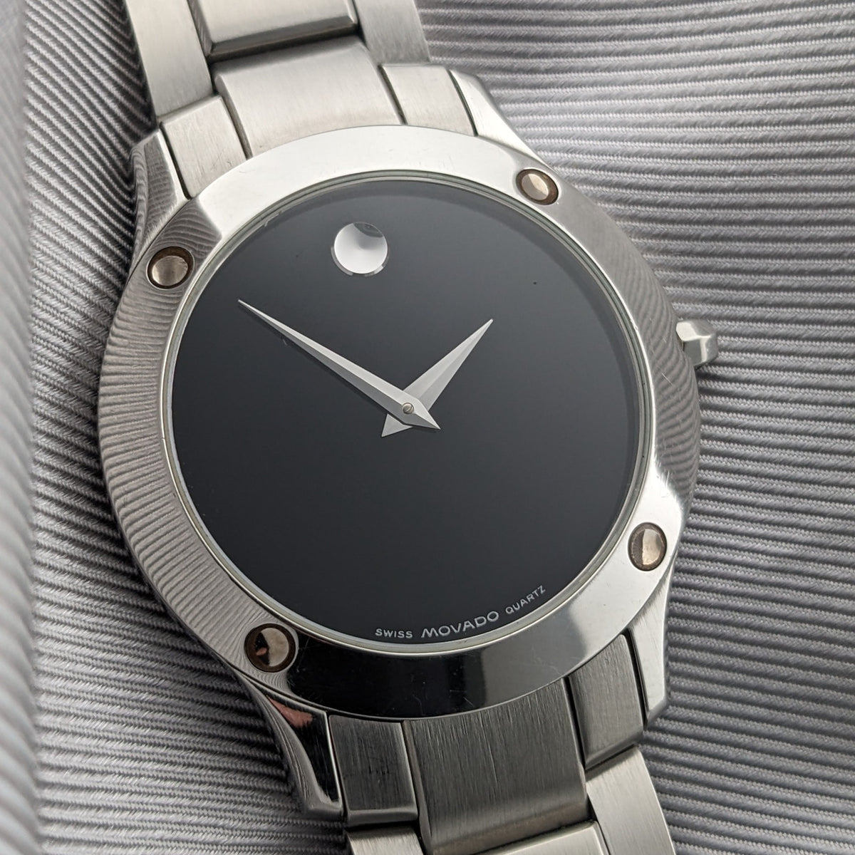 Cadran de montre Movado incliné montrant le cadran noir, aiguilles argentées, lunette polie, attaches de lunette rondes exposées et bracelet.