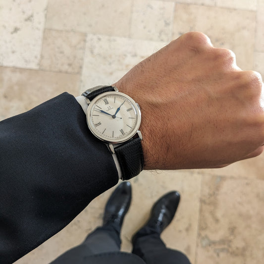 Wider wrist photograph of the Omega watch with its cream dial, blue hands and black strap, viewed above polished shoes.