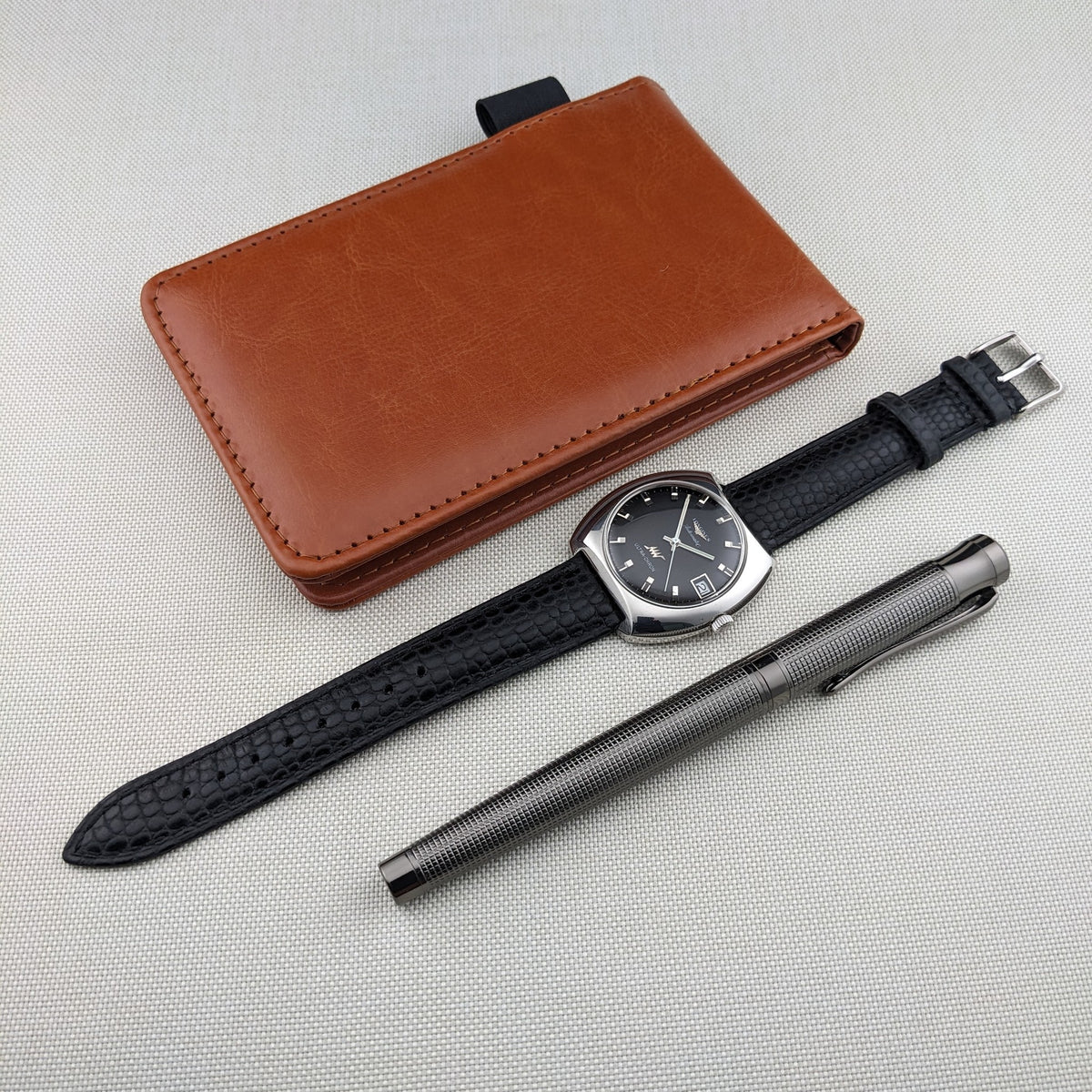 Longines Ultrachron reference 8429 arranged with a brown leather wallet, black strap, and textured silver pen on a light fabric surface.