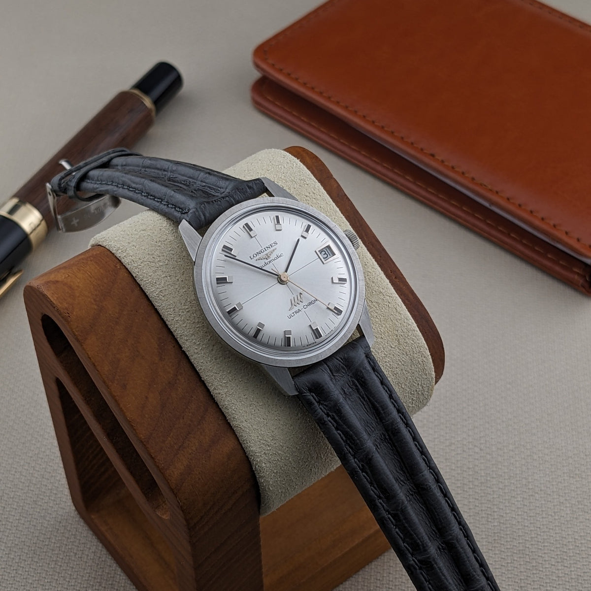 Longines Ultra-Chron 7827 resting on a wooden watch stand, with its silver dial and black strap framed by a leather case and pen.