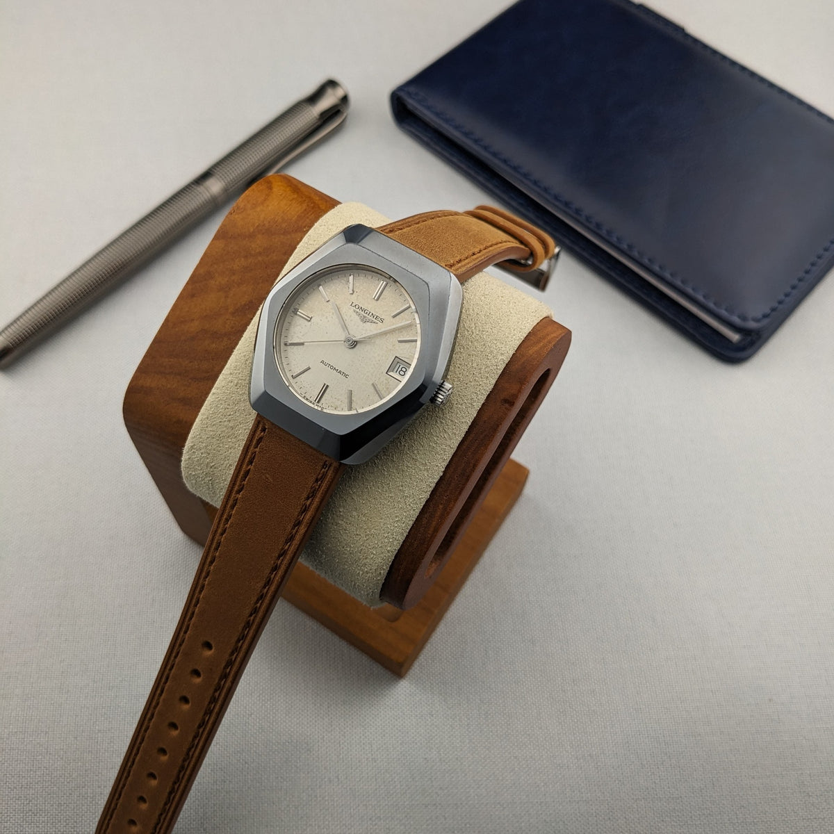 LONGINES Automatic reference 3042-633 resting on a wooden watch stand, with its cream dial, brown strap, pen and blue case visible.