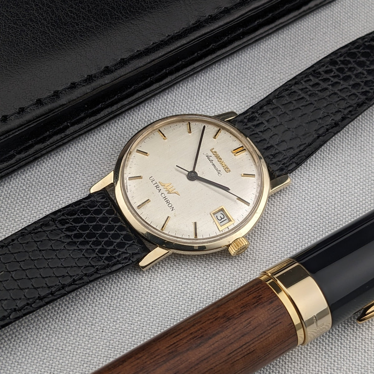 Angled composition of the Longines Ultra-Chron reference 69-556AC on fabric beside a black case and wood-and-black pen.