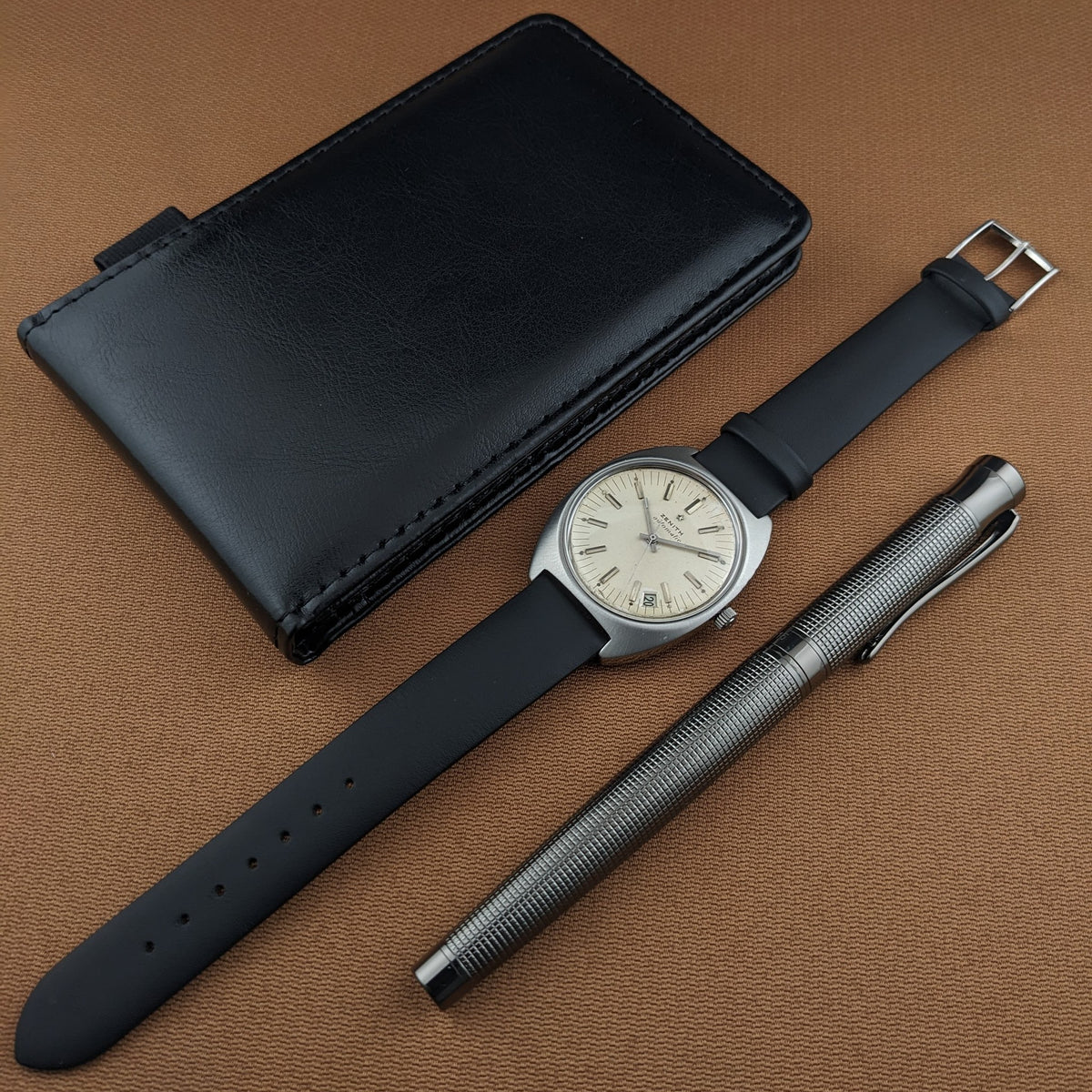 Zenith Automatic reference GR 720001 arranged with a black case and textured silver pen on brown fabric.