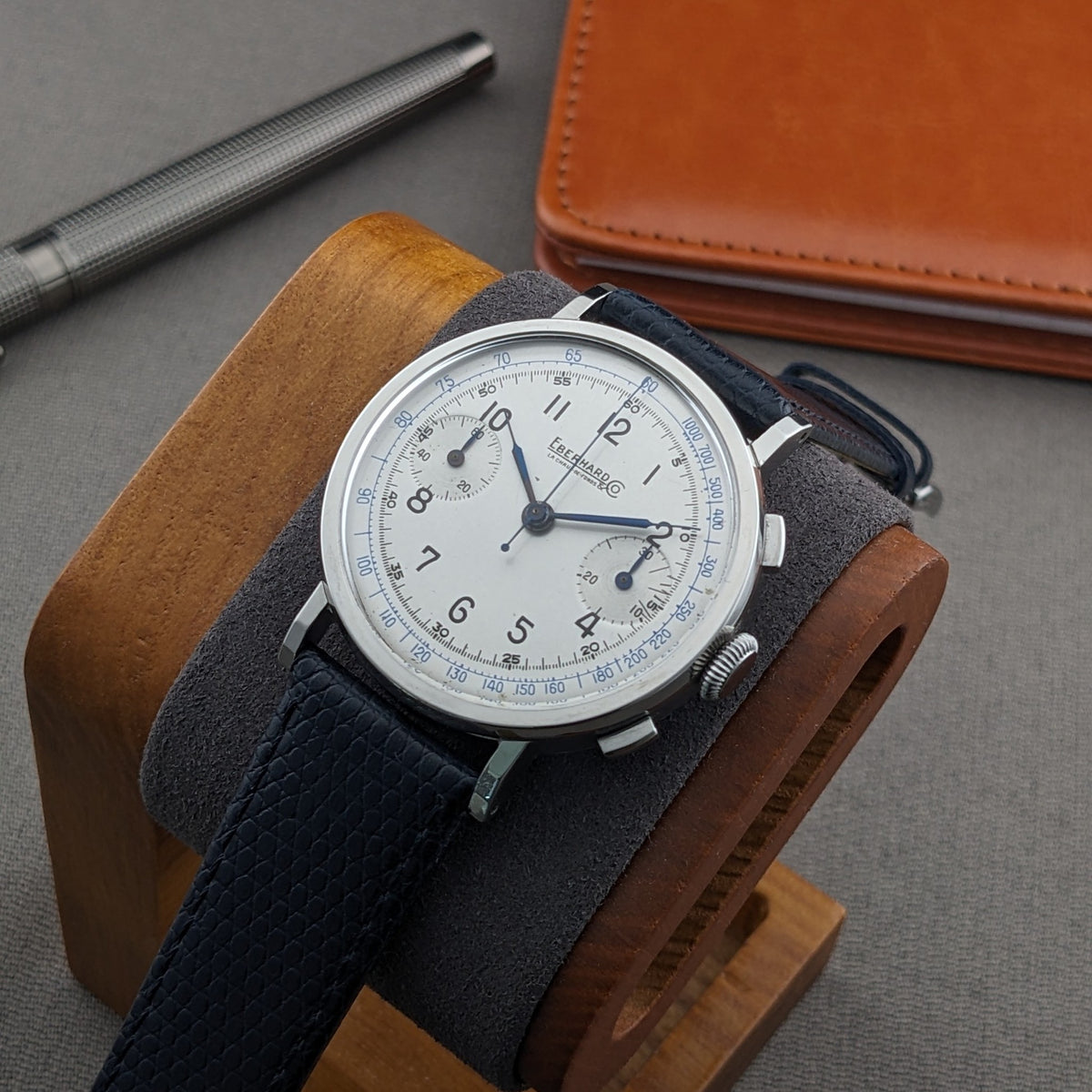 Eberhard & Co.. Pre-Extra Fort rests on a wooden watch stand, showing its cream chronograph dial, blue strap and surrounding wallet.