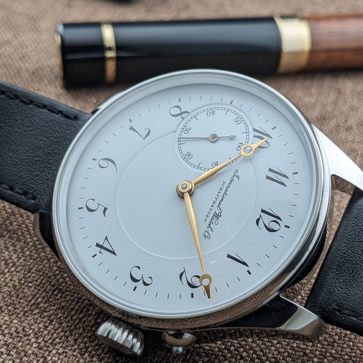 IWC Schaffhausen सफेद डायल का क्लोज़-अप, स्टाइलाइज्ड अरबी अंक, सुनहरे हाथ, मिनट ट्रैक और छोटे सेकंड्स रजिस्टर के साथ।