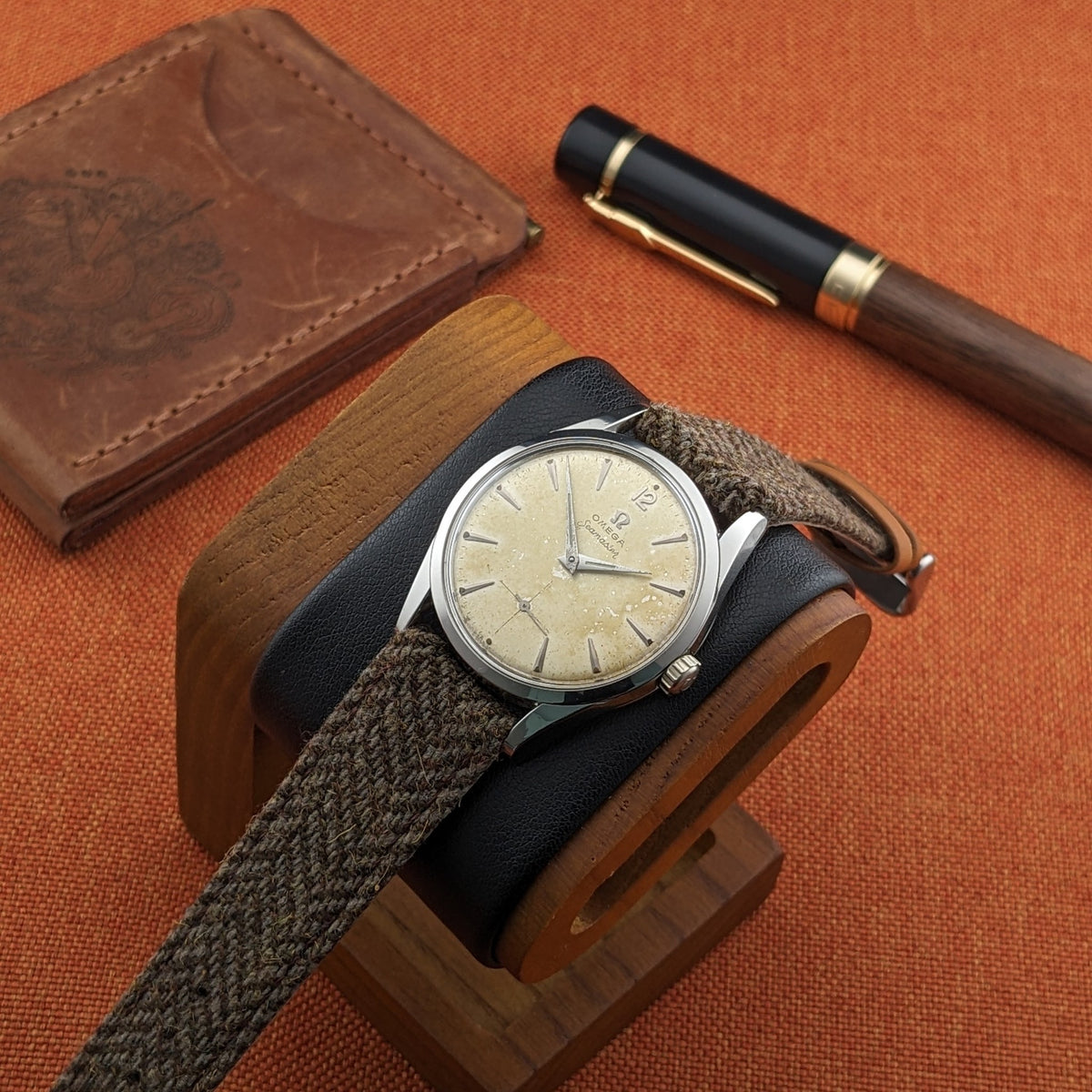 Omega Seamaster reference 29374 displayed on a wooden stand with its woven strap, leather case and black-and-wood pen in the background.