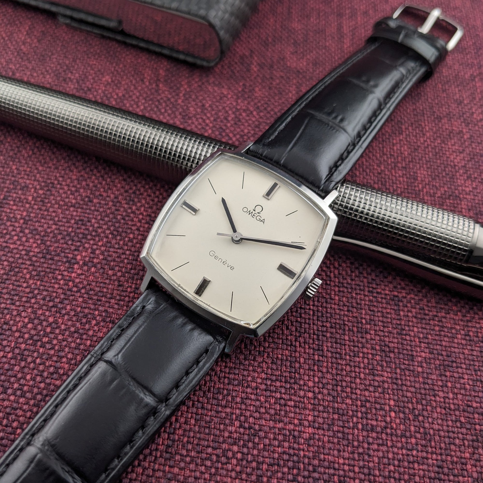 Omega Genève reference 131.022 positioned across a silver pen, with its cream dial, black crocodile-pattern strap, and buckle visible.