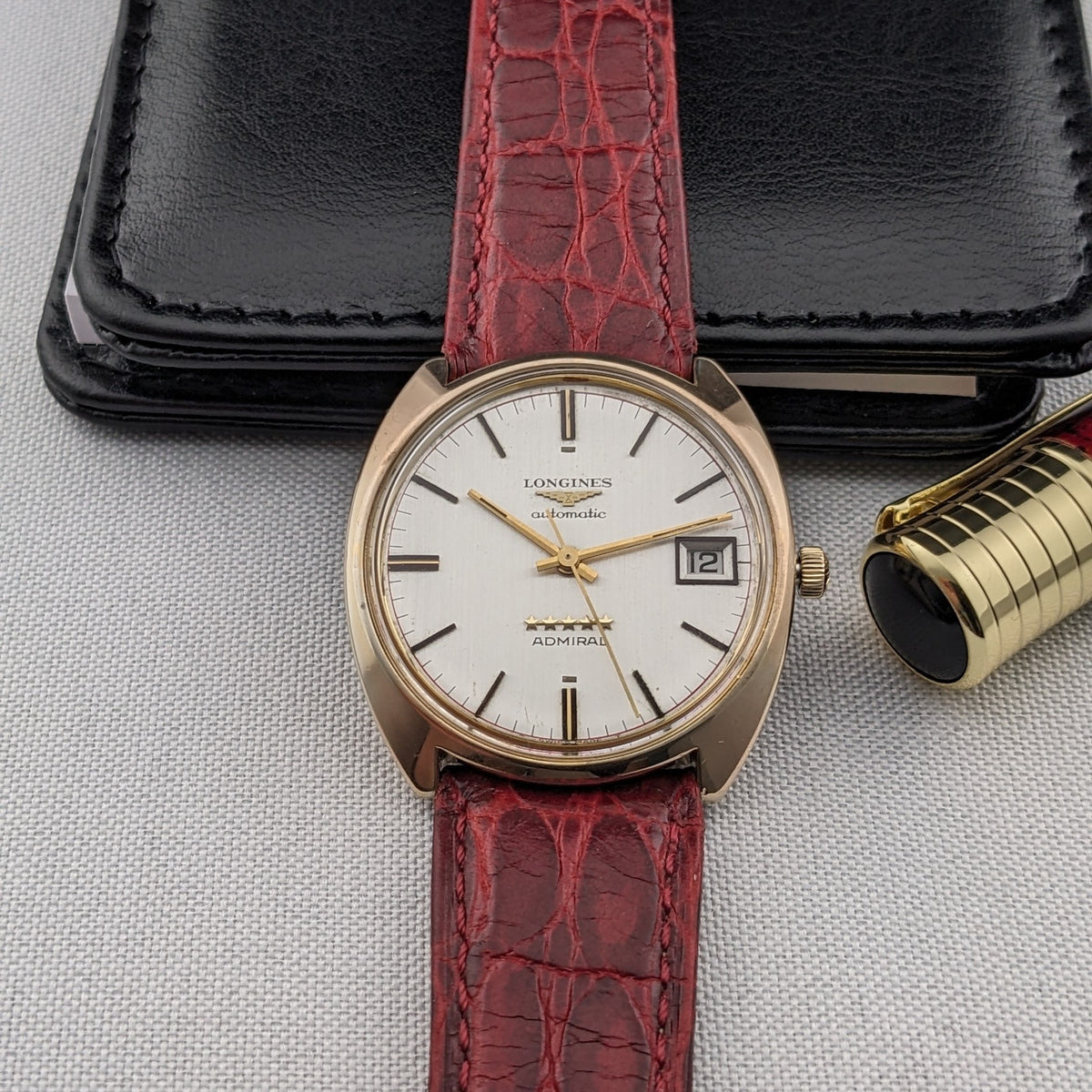 Draufsicht der Longines Admiral Referenz 8343-1 auf einer strukturierten Oberfläche, neben einem schwarzen Etui und einem goldfarbenen Stift.