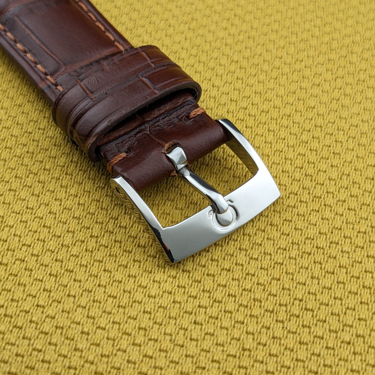 Omega Jumbo Referenz 2505-10 braunes Lederarmband endet in einer polierten Metallschließe auf einer strukturierten goldenen Oberfläche.