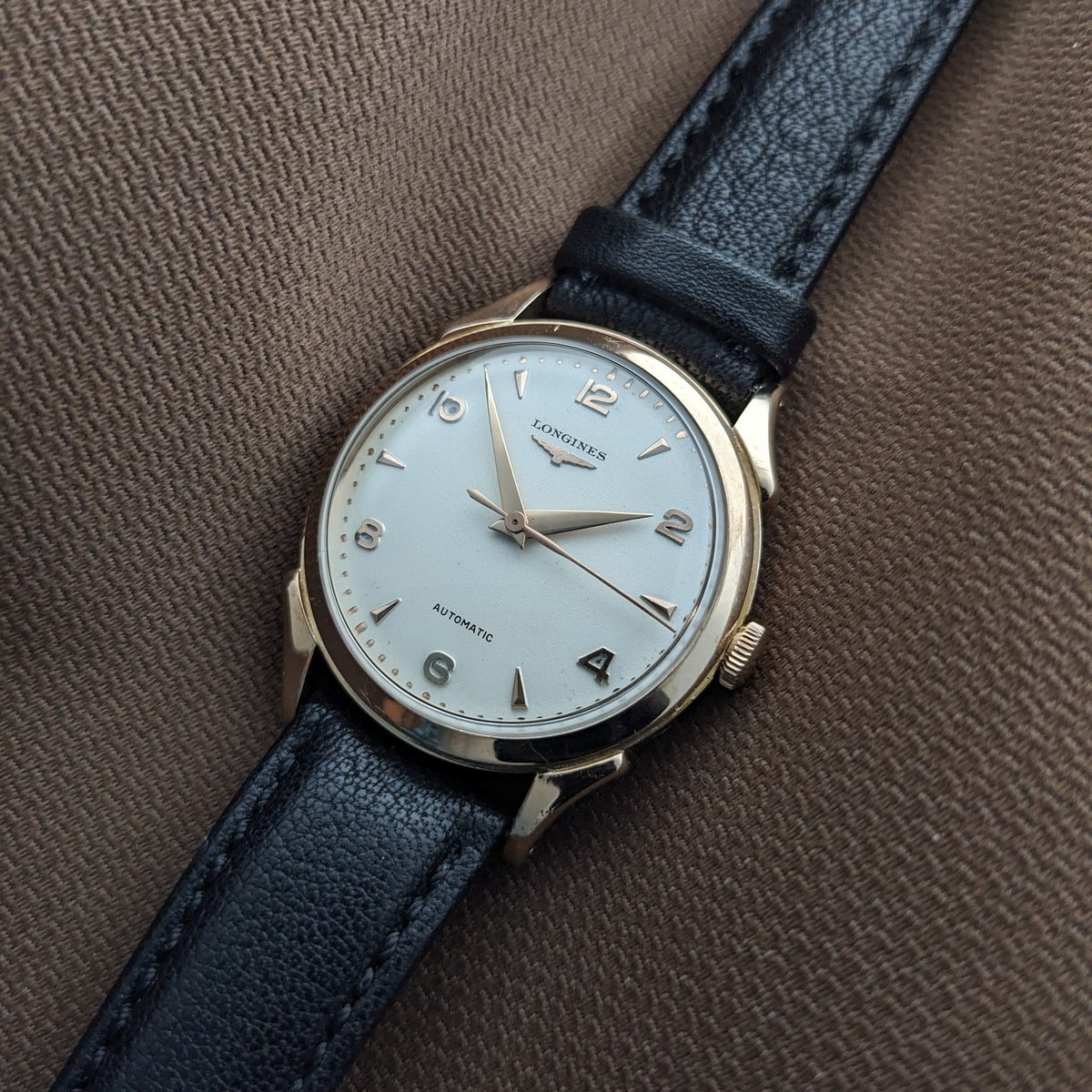 Longines Automatik Referenz 6058 auf braunem Stoff, mit hellem Zifferblatt, applizierten Ziffern, goldfarbenem Gehäuse und schwarzem Armband.