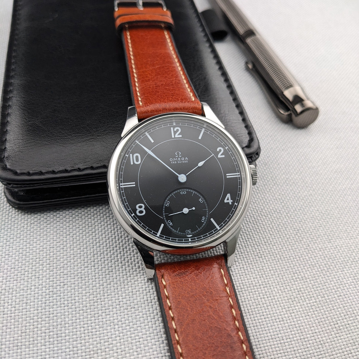 Omega-Armbanduhr mit dunklem Zifferblatt und braunem genähtem Lederarmband neben einem schwarzen Etui und silbernem Stift auf einer strukturierten Oberfläche.