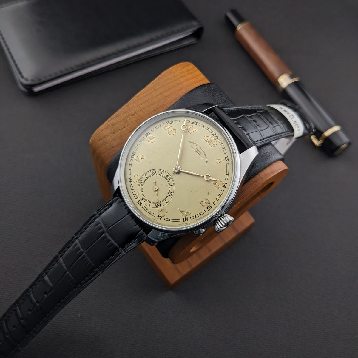 Deutsche Präzisions-Uhrenfabrik Glashütte Armbanduhr auf einem Holzständer, begleitet von einer schwarzen Geldbörse und einem Holz-und-schwarzen Stift.