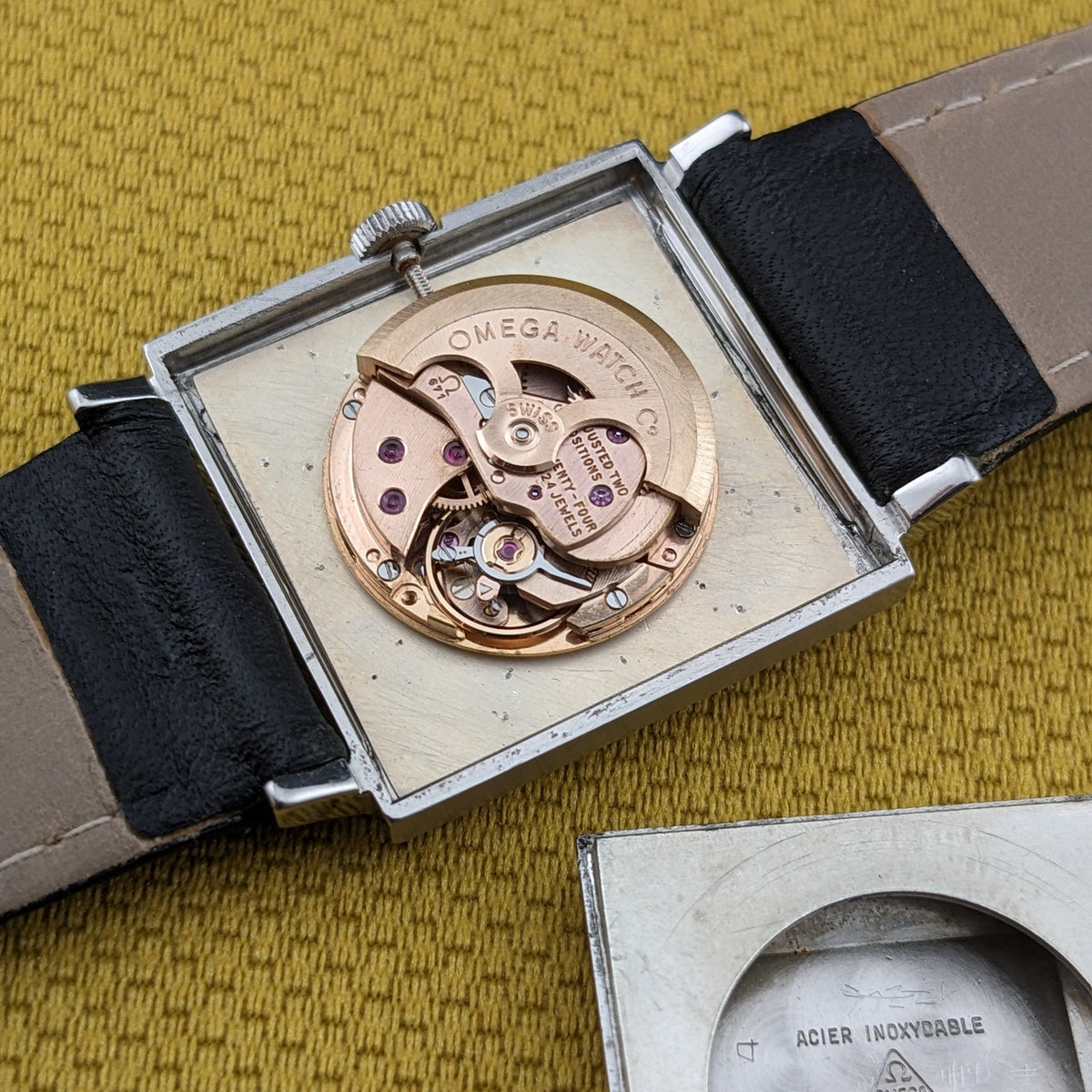 Omega De Ville Automatik 161.014 geöffnet, zeigt rundes mechanisches Uhrwerk im quadratischen Gehäuse, mit ausgezogenem Armband auf senffarbenem Stoff.