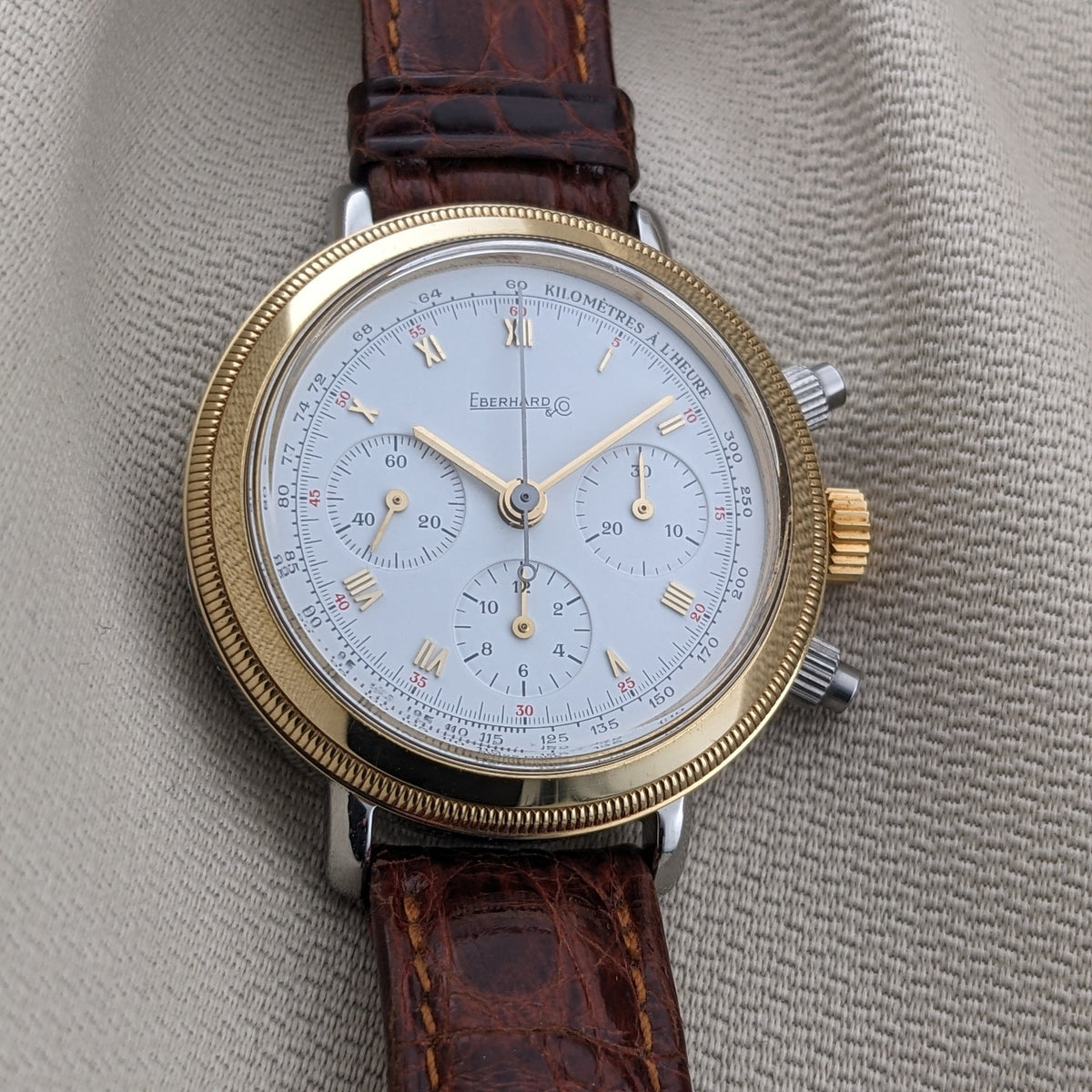 Front-facing Eberhard & Co. Godron Chrono reference 32031 on textured fabric, with a white multi-register dial, fluted bezel and brown strap.
