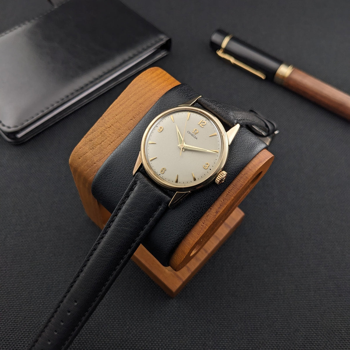 Omega Classic référence 14392-61-SC repose sur un support en bois, avec son cadran crème et son bracelet noir visibles à côté d'un étui et d'un stylo.