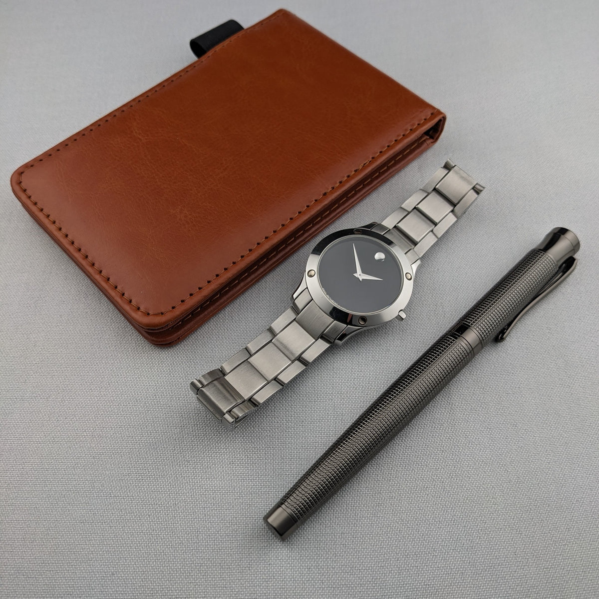 Montre Movado disposée à côté d'un étui en cuir brun et d'un stylo texturé foncé sur une surface en tissu clair.