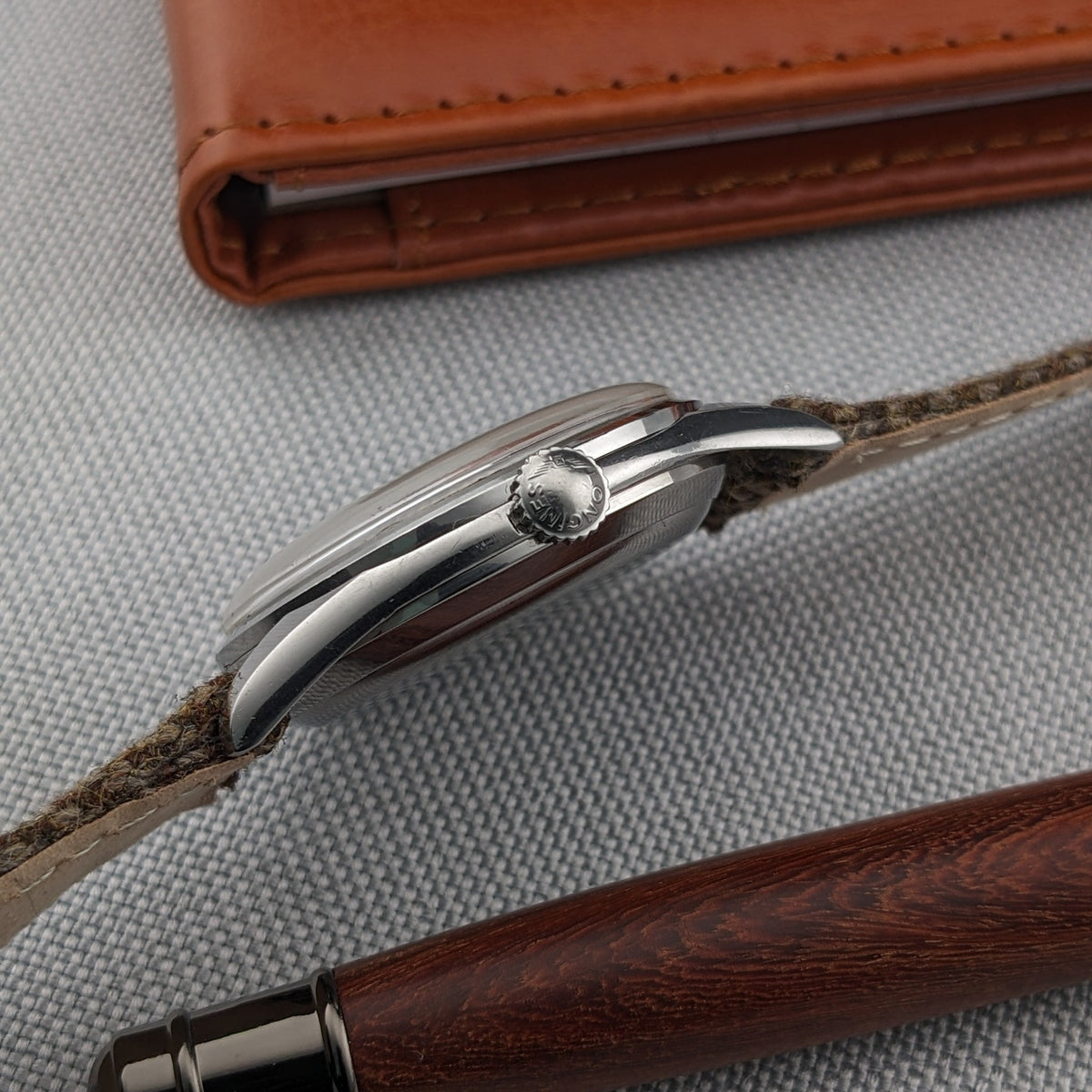 Side profile of the Longines Classic reference 8135-5 showing the curved case, crown and brown woven strap beside a leather wallet and pen.