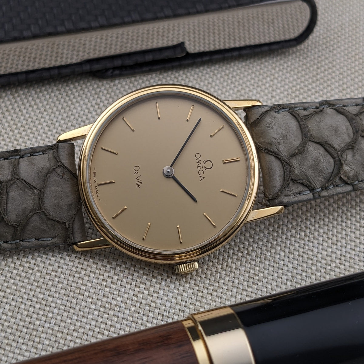 Angled Omega De Ville reference 111 0156 showing the cream dial, gold-tone bezel, crown, blue hands, and gray strap.